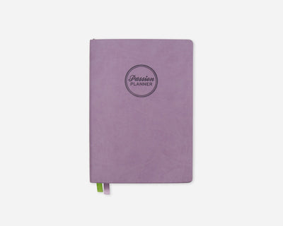 A lavender purple planner with a circular debossed logo reading 'Passion Planner' on the front, shown on a clean white background with a small green and purple ribbon bookmark peeking out from the bottom.
