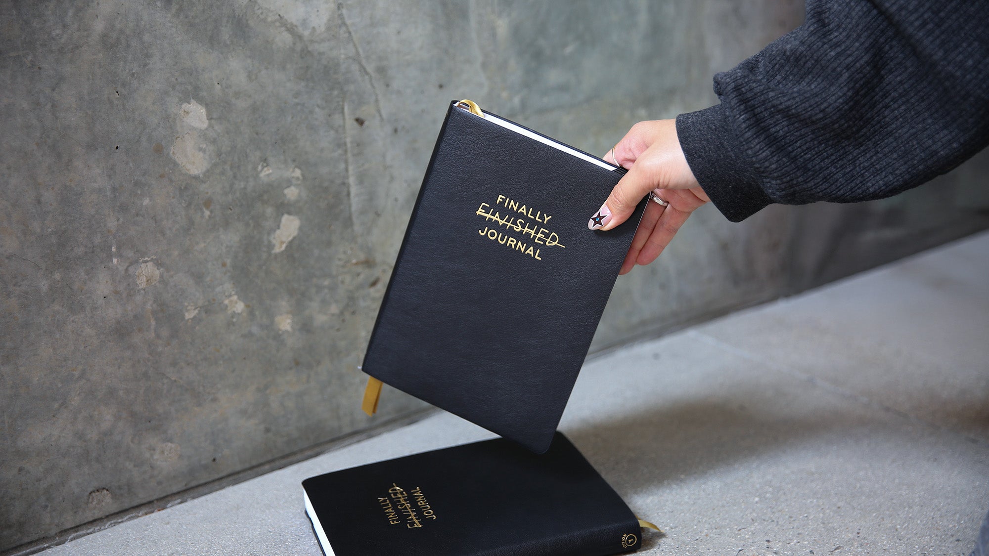 Person holding a black journal titled 'Finally Finished Journal' against a concrete wall.