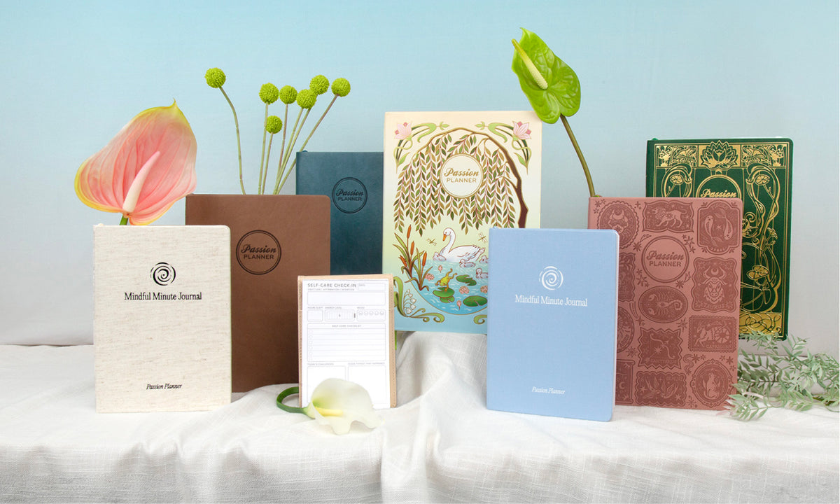 A collection of Passion Planner journals and 2027 planners in various colors and textures, arranged on a white draped tablecloth with decorative flowers and greenery against a light blue background.