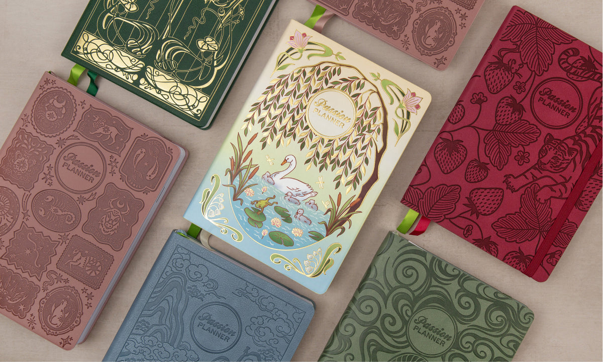 Six decorative planner  covers arranged in a flat lay on a light neutral background. The covers feature embossed floral and decorative patterns in muted tones including dusty rose, forest green, coral red, light blue, and olive green, with one central cover showing a whimsical tree-of-life illustration with a swan and pond.