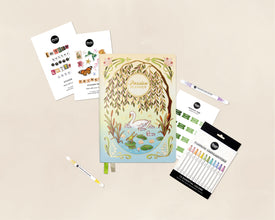 A stationery flat lay featuring a pastel illustrated planner titled 'Passion Planner' with a willow tree and swan design, surrounded by sticker sheets, two white pens, and a pack of colorful highlighter pens on a light beige background.