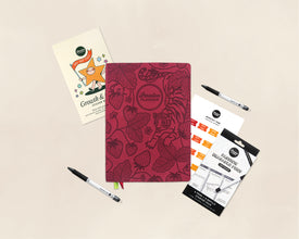 A styled flat-lay of a 'Passion Planner' embossed planner in deep red with botanical line art, surrounded by stationery accessories including a Growth & Goals Sticker Book, monthly tabs, and two black pens on a light beige background.