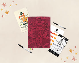 A styled flat-lay of stationery items on a light beige background with small star illustrations. The main product is a red planner with botanical strawberry and leaf patterns, labeled 'Passion Planner'.