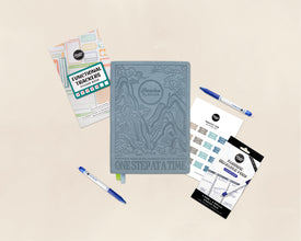 A styled flat-lay of a blue-gray embossed planner notebook in the center, surrounded by coordinating planner accessories including functional tracker stickers, blue pens, and monthly tabs on a warm beige background.