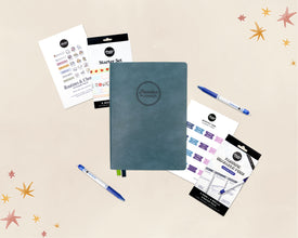 A styled flat-lay of a planner starter set on a warm beige background with decorative star graphics. The set includes a dark teal-blue planner notebook with an embossed logo, blue pens, sticker sheets, washi tape, and monthly tab stickers.
