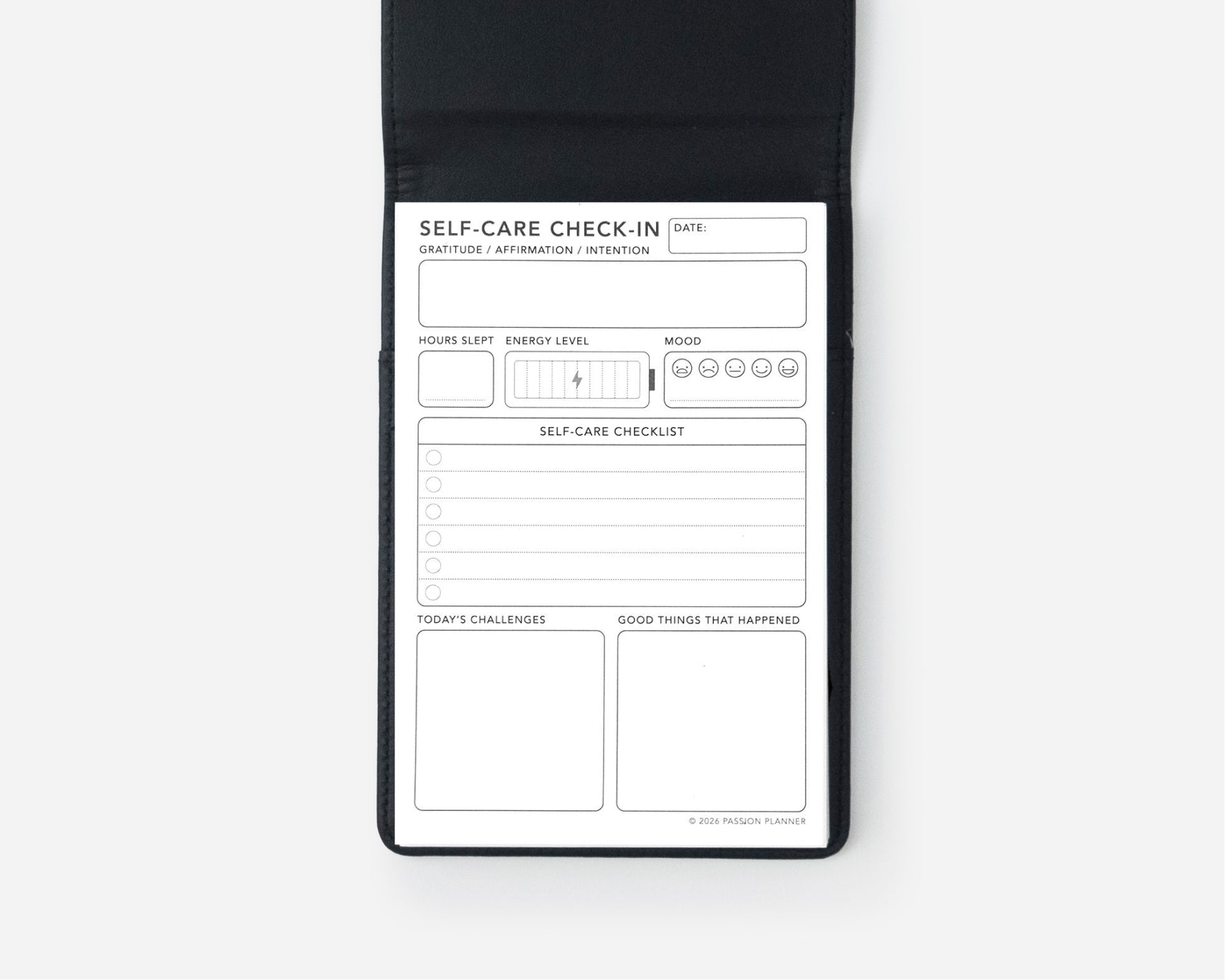 A black self-care checklist notepad shown open against a light gray background, featuring a white insert with fields for date, self-care check-in, sleep, energy level, mood, challenges, and good things that happened.