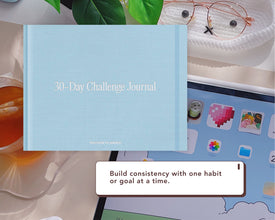 A light blue 30-Day Challenge Journal by Passion Planner is shown with a styled desk setting with a tablet, glasses, tea, and green leaves. A text bubble reads, 'Build consistency with one habit or goal at a time.'.