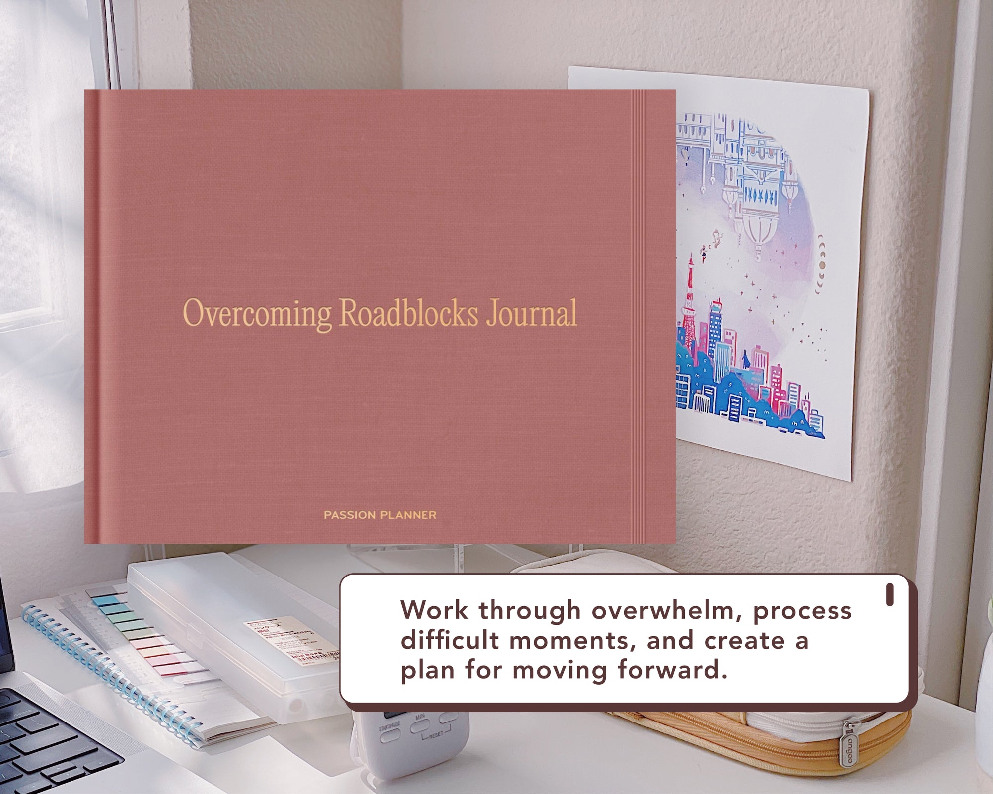 A rose pink hardcover journal titled 'Overcoming Roadblocks Journal' with gold lettering, shown on a desk alongside stationery items and a framed art print. A white text box overlay reads: 'Work through overwhelm, process difficult moments, and create a plan for moving forward.'