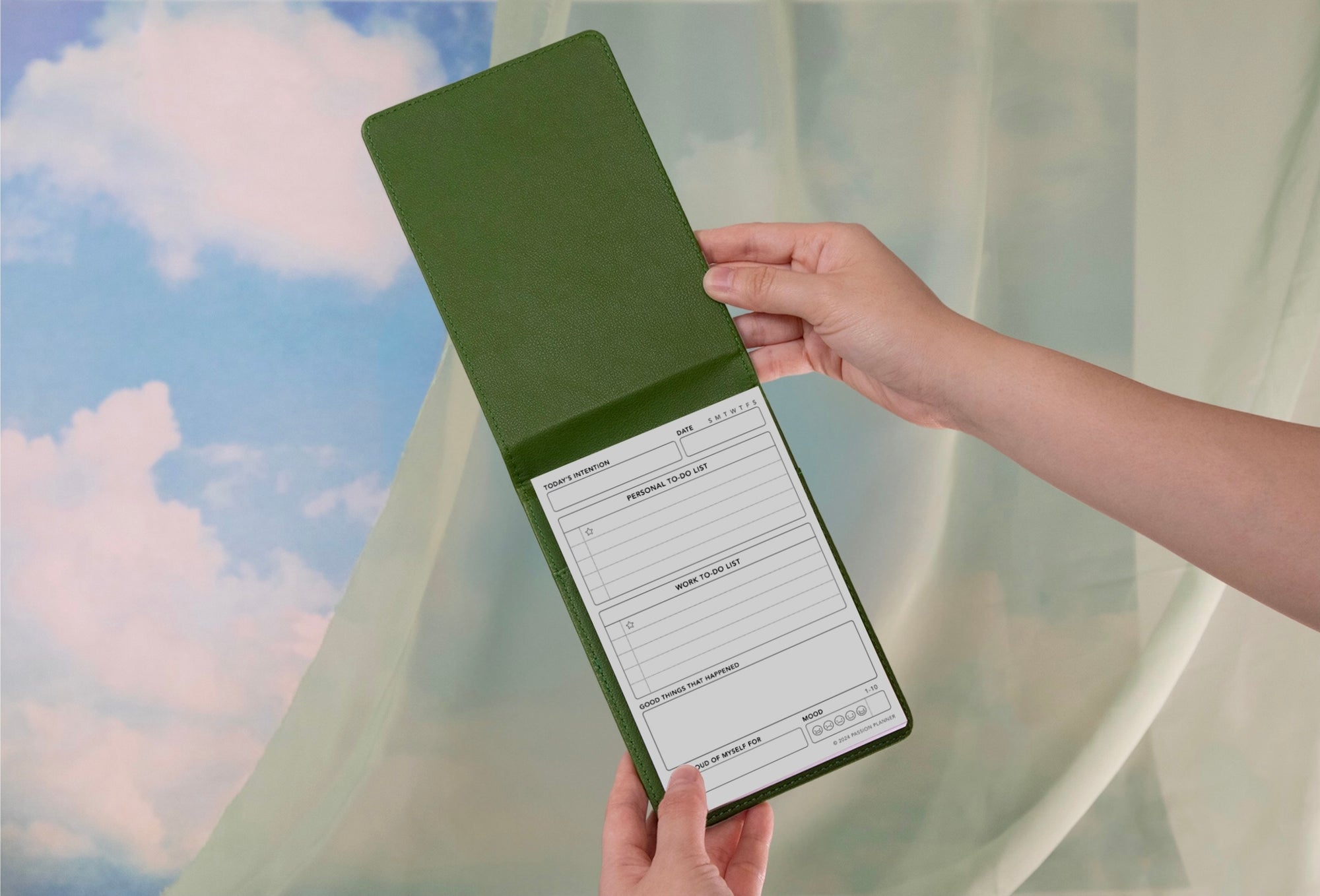 Hand holding a green notepad with a daily schedule and to-do list with curtains and clouds in the background.
