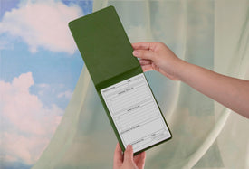 Hand holding a green notepad with a daily schedule and to-do list with curtains and clouds in the background.