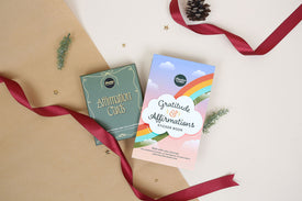 Affirmation Cards and 'Gratitude & Affirmations' Sticker Book on a beige surface with a red ribbon and pine cone.