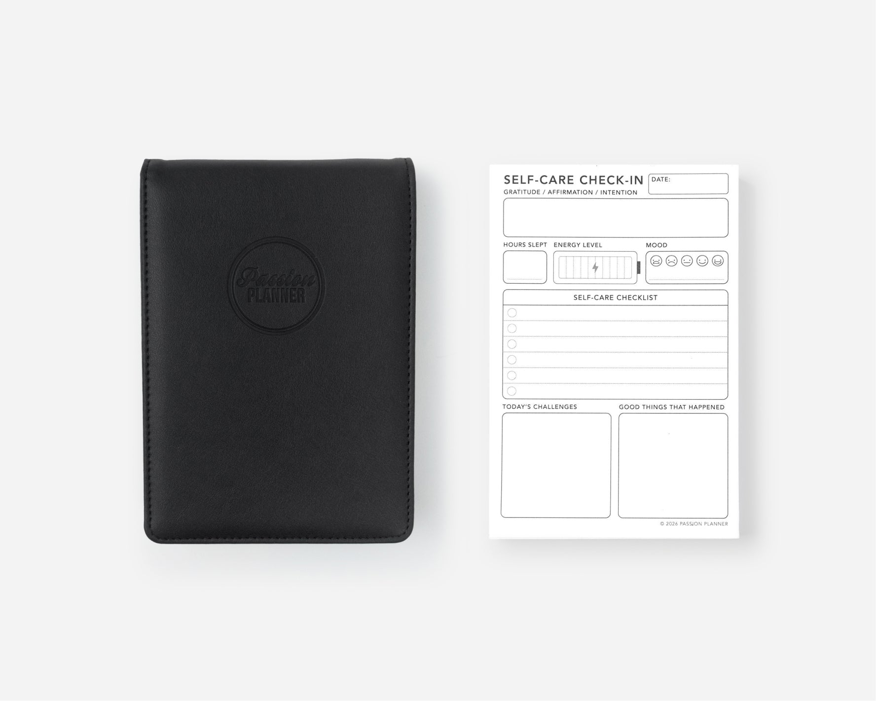 A black leather self-care notebook cover with an embossed circular logo next to a white 'Self-Care Check-In' notepad on a light gray background.