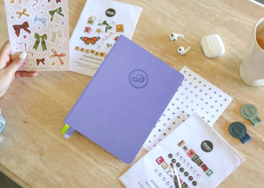 Purple journal with a logo on a wooden table with stickers and a mug.