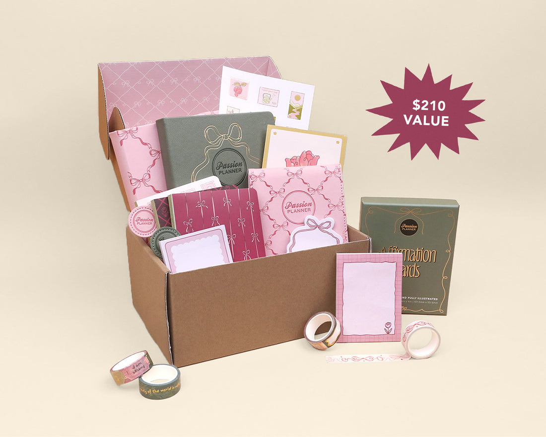 A pink-themed stationery gift set displayed in an open brown cardboard box, with matching boxed items placed beside it on a warm beige background. A magenta starburst in the upper right reads &